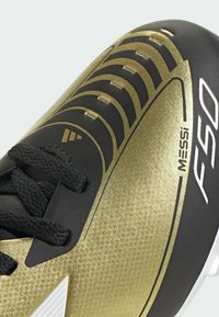Botas de fútbol negras y doradas con parte superior texturizada, que cuentan con un cierre de cordones y acentos blancos. El diseño es aerodinámico con notables patrones dorados.
