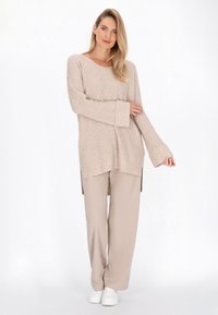 Maglione beige a maglia con vestibilità morbida, maniche lunghe e spacchi laterali, abbinato a pantaloni beige a gamba larga e sneakers bianche.