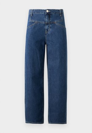 Frau trägt einen braun-blau karierten Blazer, schwarzes Hemd, weißes Unterhemd, lockere blaue Jeans, schwarze Socken und marineblaue Sneakers, steht vor weißem Hintergrund.