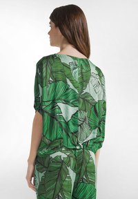 Grøn bluse med bladmønster lavet af letvægtsstof, der har en løs pasform, sænkede skuldre og en rynket ryg.