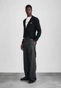 Black cardigan with a fox emblem, worn over a white T-shirt. Paired with gray pleated trousers and black shoes. Smooth texture, relaxed fit.