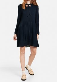 Marinblå klänning med veck och långa ärmar samt knytning vid halsen. Buren med ljusa strumpor och beige loafers med mörka sulor.