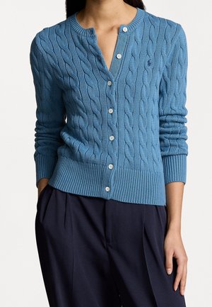 Personne portant un cardigan bleu à torsades boutonné avec des boutons blancs et un pantalon bleu marine foncé, debout avec une main dans la poche.