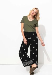 T-shirt vert olive à col en V, pantalon ample noir avec des motifs brodés argentés et un ourlet décoratif. Chaussures brunes à enfiler. Fond blanc.