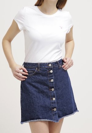 Frau trägt ein weißes kurzärmeliges T-Shirt, das in einen hoch taillierten blauen Jeansrock mit Knopfleiste und ausgefranstem Saum gesteckt ist.