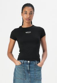 Melnas īsām piedurknēm t-krekls ar pieguļošu dizainu, kuram uz krūtīm ir balts "TOMMY JEANS" logo. Mīksts kokvilnas materiāls.