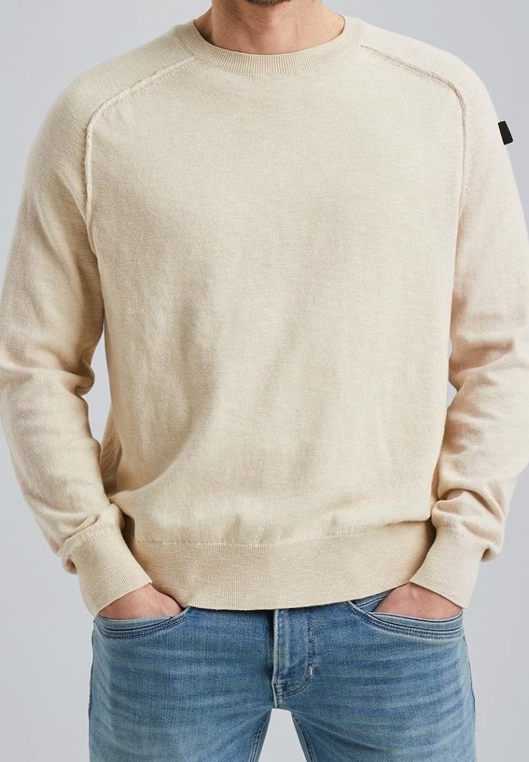 Beige gebreide trui met geribde ronde hals, raglanmouwen, gestructureerde afwerking en subtiel logodetail op de linkerschouder. Draagt blauwe spijkerbroek.