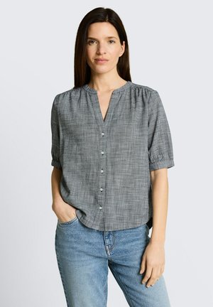 Vrouw met lang donker haar die een grijs geruit blouse met korte mouwen en knopen draagt en een blauwe spijkerbroek, staand met één hand in de zak.