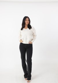 Maglione bianco a collo a V con piccoli motivi a cuore beige, abbinato a jeans neri aderenti. Texture liscia, vestibilità rilassata e design minimalista.