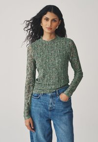 Green long-sleeve top with smocked texture and floral print on sheer sleeves, paired with blue high-waisted jeans.