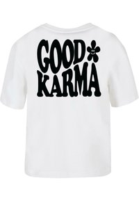 T-shirt en coton blanc avec le texte noir en gras "BONNE KARMA" et un petit graphique de fleur souriante au dos. Manches courtes, coupe décontractée.