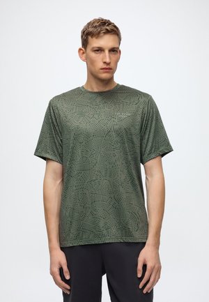 Jeune homme portant une chemise à manches courtes verte à motifs et un pantalon noir, debout devant un fond blanc uni.