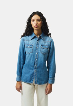 Donna con capelli ricci che indossa una camicia di jeans blu abbottonata con tasche sul petto e pantaloni color crema su uno sfondo semplice.