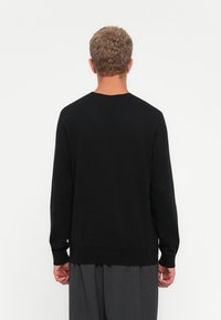 Homme aux cheveux courts et bouclés portant un pull noir à manches longues et un pantalon gris, vu de dos sur un fond blanc uni.