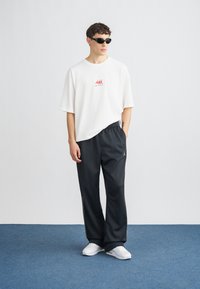 Biele nadrozmerné tričko s červenou grafikou, skombinované s čiernymi voľnými teplákmi a bielymi teniskami. Doplnky zahŕňajú čierne slnečné okuliare.