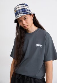 Pelēks T-krekls ar "Dickies" logo uz krūšu, kopā ar zilu un baltu rūtainu vāciņu. Garas tumšas matis. Melni bikses.