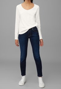 Vrouw die een witte top met lange mouwen draagt, donkerblauwe strakke jeans en witte hoge sneakers, staand tegen een effen grijze achtergrond.