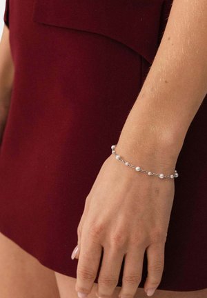 Hand trägt ein filigranes silbernes Armband mit kleinen Perlen, ruht neben dem oberen Oberschenkel, der in dunkelrotem Stoff gekleidet ist.