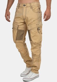Pantalons cargo beiges avec plusieurs poches, couture noire, accents côtelés sur les jambes et une coupe ajustée. Stylés avec des baskets blanches.