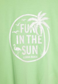 Hellgrünes T-Shirt mit weißem, rundem Grafikdesign, auf dem "SPASS IN DER SONNE", eine Palme, ein Flamingo und fliegende Vögel abgebildet sind. Weicher Stoff.