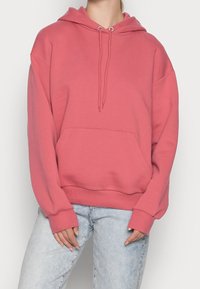 Růžová mikina s kapucí, přední kapsou, dlouhými rukávy a stahovacími šňůrkami, doplněná světle modrými džínami. Měkký materiál a volný střih.