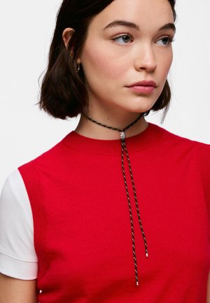 Junge Frau mit kurzen dunklen Haaren, die ein rotes Top mit weißen kurzen Ärmeln und eine schwarze Kordelhalskette mit einer weißen Perle trägt.