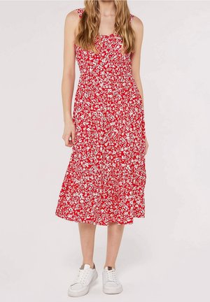 Femme portant une robe midi rouge sans manches avec imprimé floral blanc et des baskets blanches, debout devant un fond uni.