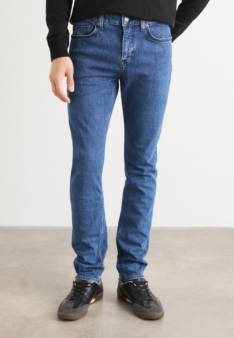 Jeans de denim azul ajustados con un diseño clásico de cinco bolsillos, costuras en contraste y cierre de botón metálico, combinados con zapatillas deportivas negras.