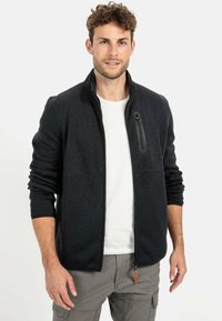 Donkergrijze fleecejack met een hoge kraag, ritssluiting en een borstzak. Witte t-shirt eronder, tegen een neutrale achtergrond.