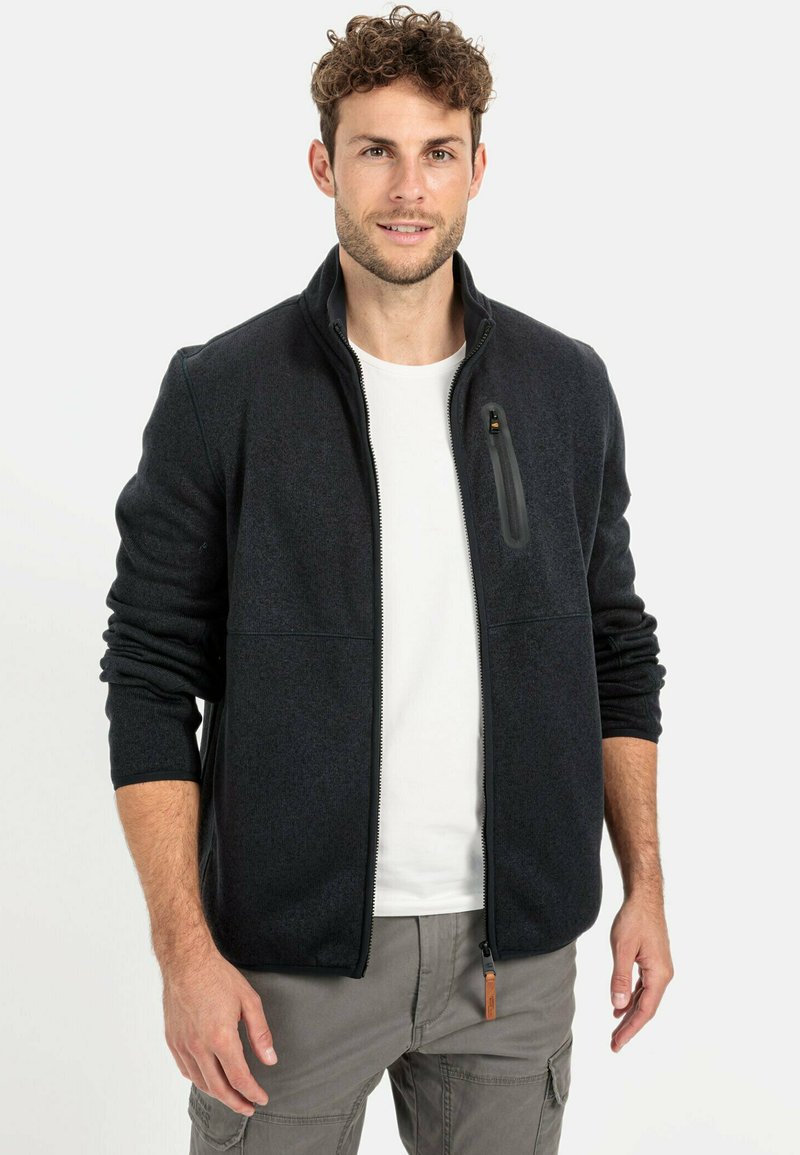 Donkergrijze fleecejack met een hoge kraag, ritssluiting en een borstzak. Witte t-shirt eronder, tegen een neutrale achtergrond.