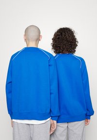 Felpa blu con maniche raglan, scollo rotondo e dettagli di cucitura bianchi. Indossata sopra pantaloni della tuta grigi, evidenziando un fit rilassato.