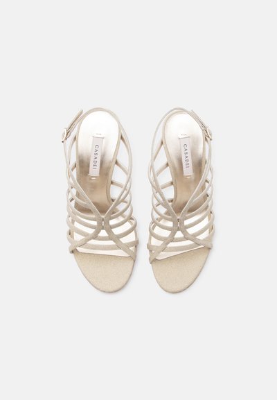 Ζευγάρι μπεζ γυναικείων σανδαλιών με λουράκια και λουράκια με πλάτη, με το λογότυπο Casadei στις εσωτερικές σόλες, εμφανιζόμενο από πάνω σε ανοιχτό φόντο.