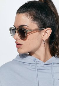 Läbipaistvad raamiga prillid, tume objektiiv, nurkadega kuju ja diskreetne bränding templil, kantud helesinise kapuutsiga.