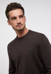 Brauner Strickpullover mit Rundhalsausschnitt und gerippten Bündchen, der eine weiche Textur und eine figurbetonte Passform aufweist. Der Hintergrund ist schlicht weiß.