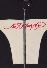 Sort og cremefarvet jakke med frontlynlås og rødt broderet "Ed Hardy"-logo henover brystet.
