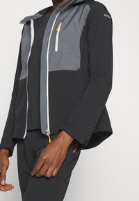 Personne portant une veste zippée noire et grise avec une tirette jaune et un pantalon noir avec un petit détail rouge sur la poche.