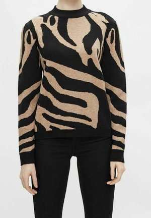 Persoon die een zwart-beige trui met een abstract patroon en lange mouwen draagt, met een zwarte broek, staand tegen een witte achtergrond.