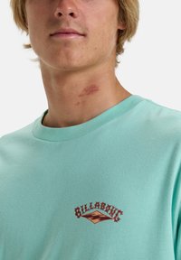 Mentolové tričko s krátkým rukávem a kulatým výstřihem, na přední straně s červeným a zlatým logem "Billabong" na hrudi. Hladká textura látky.