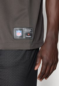 NFL branded siva majica kratkih rukava s teksturiranim završetkom i crnom etiketom koja sadrži logo i tekst, uparenim s crnim šortsevima s tufnicama.
