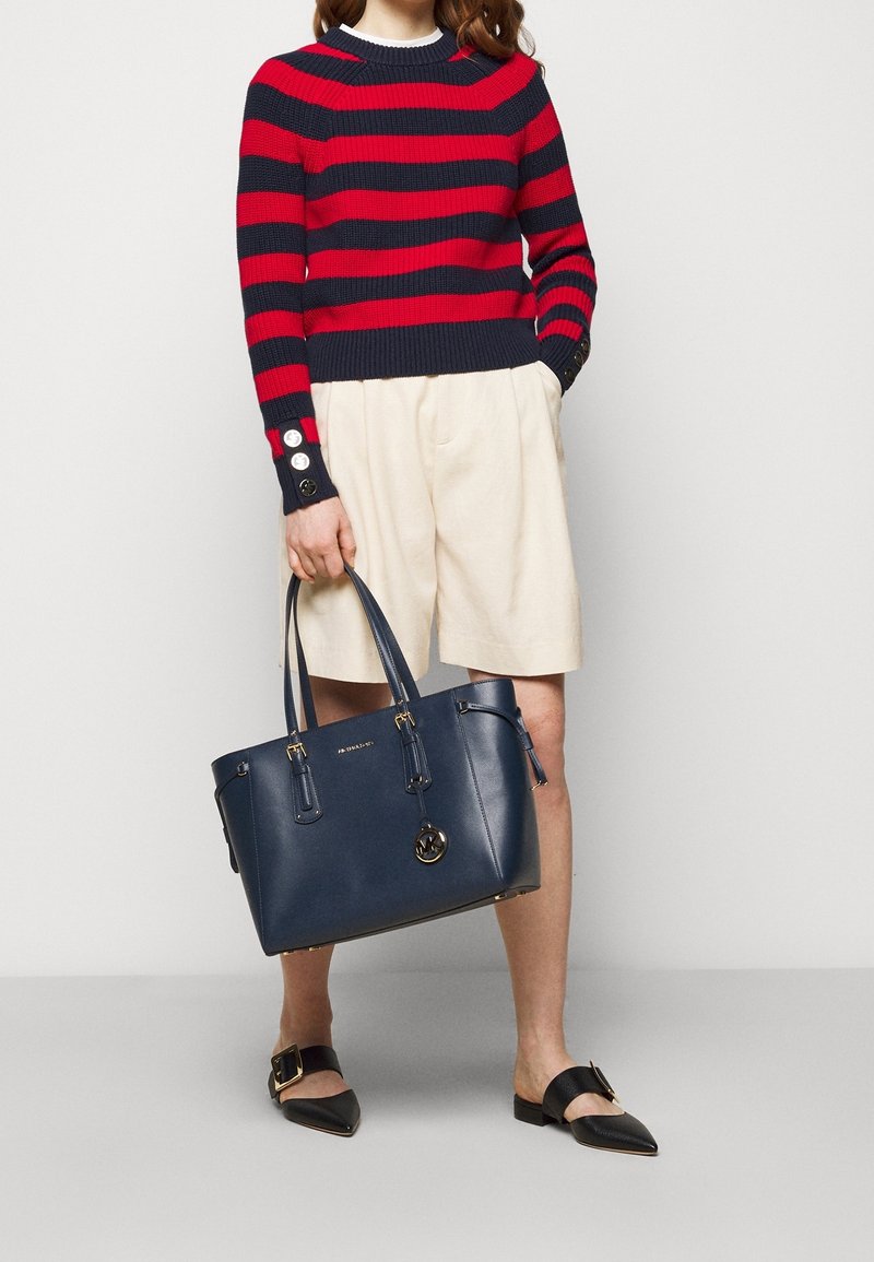Personne portant un pull rayé rouge et marine, un short crème, des chaussures noires pointues avec boucles, tenant un grand sac à main marine avec des accents dorés.