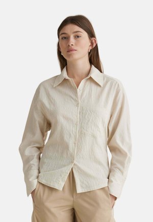 Mujer con camisa beige de rayas abotonada con cuello y pantalones marrón claro, de pie con las manos en los bolsillos sobre un fondo liso.