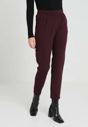 Vrouw draagt hooggetailleerde, taps toelopende donkerpaarse broek met voorplooien, zwarte geribde trui en zwarte lakleren enkellaarzen met blokhakken.