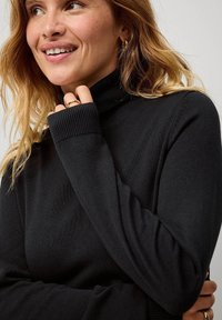 Maglione nero a collo alto realizzato in materiale a coste, con maniche lunghe e un design aderente. Mani incrociate vicino all'area del collo, in evidenza anelli.