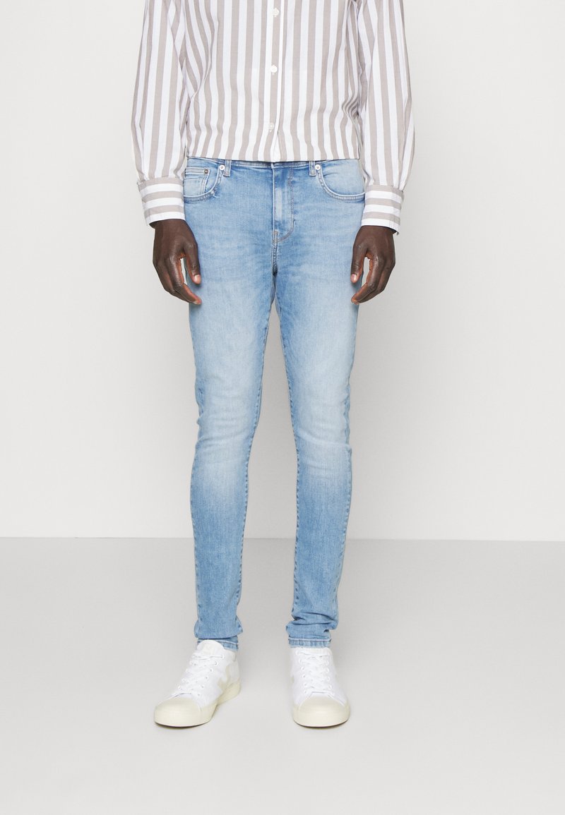 Jeans ajustados de color azul claro en tela de mezclilla, con cinco bolsillos y un ligero desvanecimiento, combinados con una camisa de rayas de manga larga y zapatillas blancas.