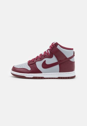 Sneaker wysokiej na kostkę z białej i ciemnoczerwonej skóry z sznurowadłami, perforowanym noskiem i ciemnoczerwonym logo Swoosh po boku, na jednolitym tle.