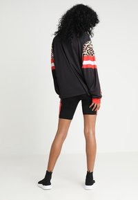 Žena stojící zády, oblečená v černém topu s dlouhým rukávem, na kterém jsou leopardí vzory a červené pruhy na rukávech, černých bikerských šortkách s červenými bočními pruhy a černých botách.