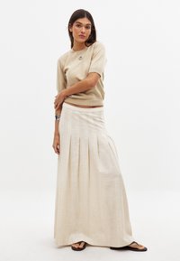 Maglietta beige a maniche corte abbinata a una lunga gonna maxi plissettata di colore chiaro. La gonna presenta una sottile texture e una vita piatta.