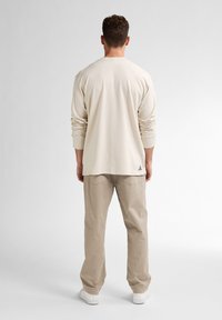 Beige langslavige shirt met een relaxte pasvorm, gecombineerd met lichtbeige broeken. Het shirt heeft een ronde hals en een subtiele logo detail aan de onderkant.
