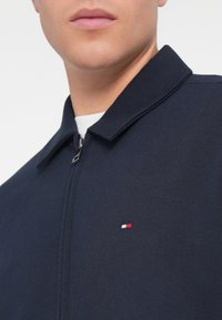 Άνδρας που φοράει ναυτικό μπλε μπουφάν με φερμουάρ, με ένα μικρό κόκκινο, λευκό και μπλε ορθογώνιο λογότυπο στο στήθος, πάνω από ένα λευκό πουκάμισο.