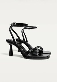 Schwarze Sandalen mit hohen Absätzen, schmalen gekreuzten Riemen, abgerundeter Zehenpartie, Knöchelriemen mit Schnalle und quadratischem Absatz. Glatte Lederstruktur.
